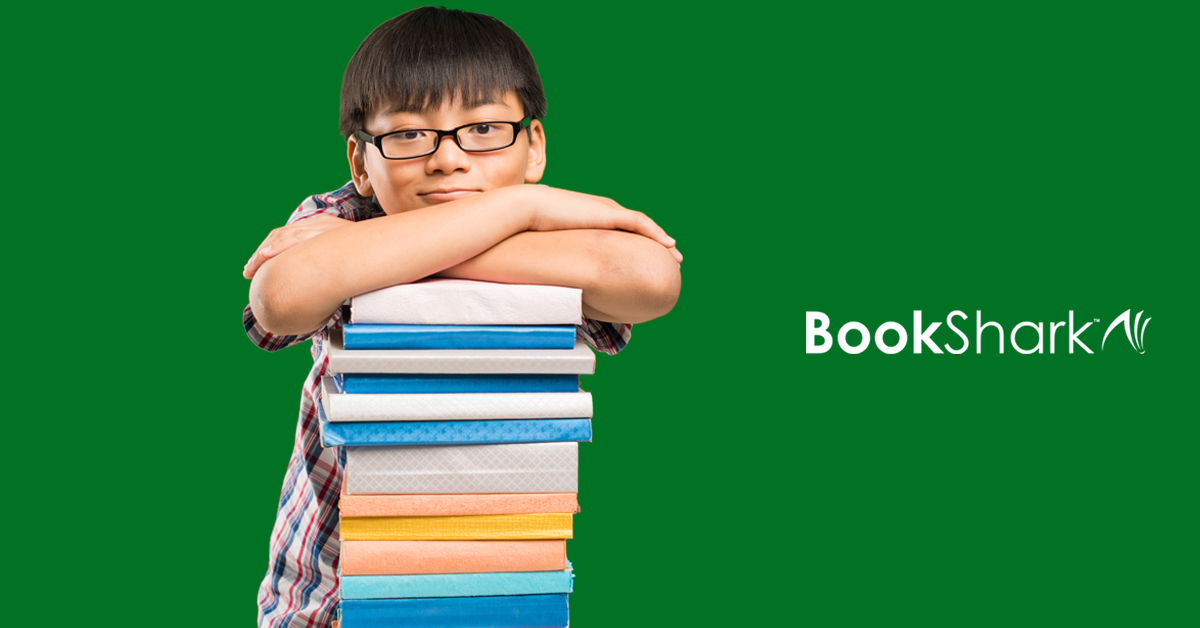 a boy wearing classes leans against a tall pile of hardback books