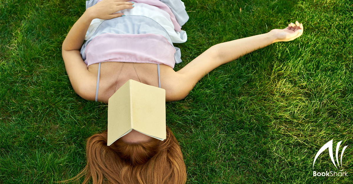 a woman in a pastel sundress lies on her back on bright green grass, an open book hiding her face