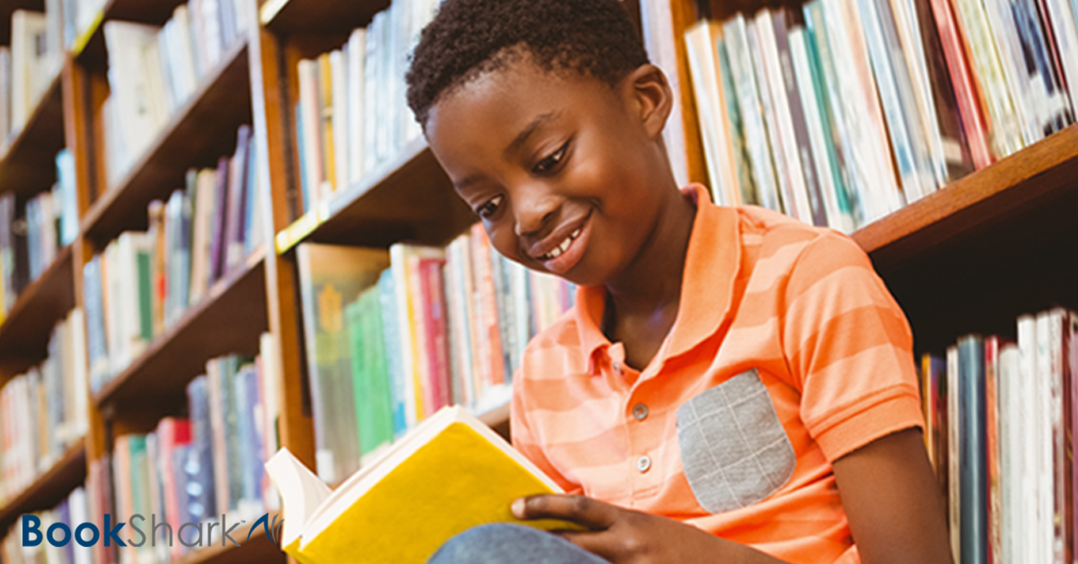 a boy sits with his back against a library bookcase, reading a book with a yellow cover
