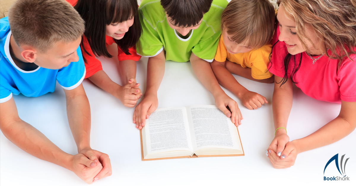 kids in vividly colored t-shirts lie prone, looking at a book