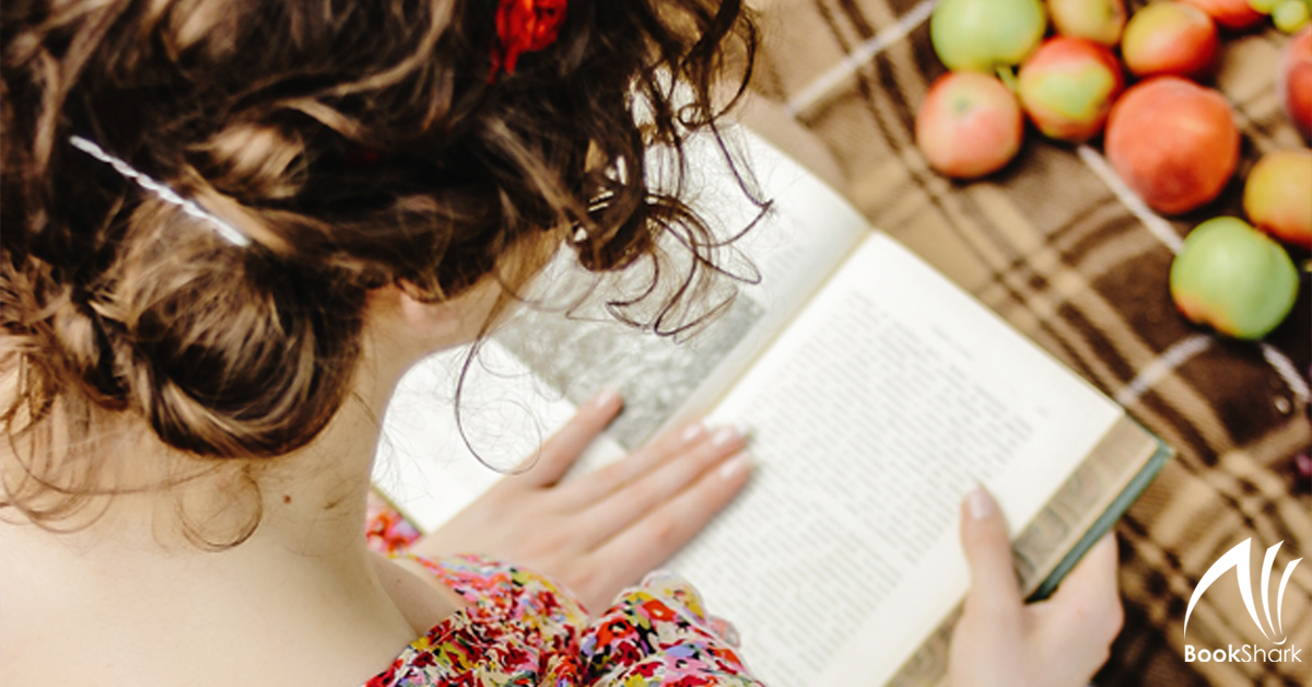 a curly haired mom in a floral dress sits on a blanket, reading a book