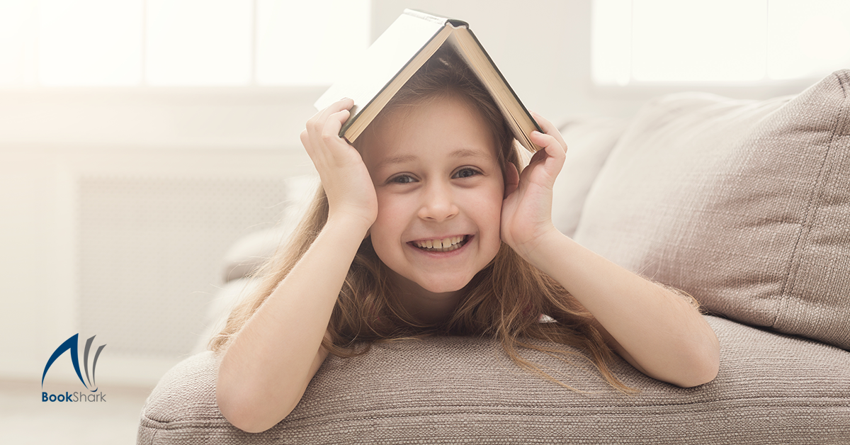 a smiling girl lies on a couch with a book on her head